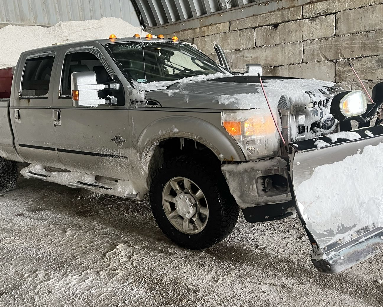 A silver pickup truck equipped with a snow plow, parked indoors on a gravel surface covered in patches of snow.