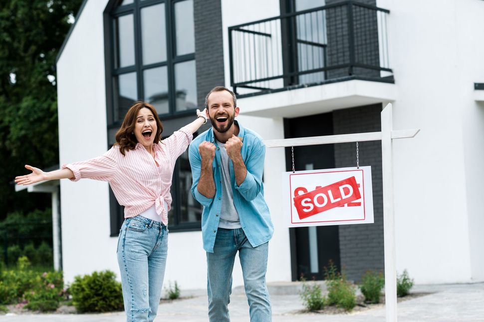 A man and a woman are standing in front of a house with a sold sign.