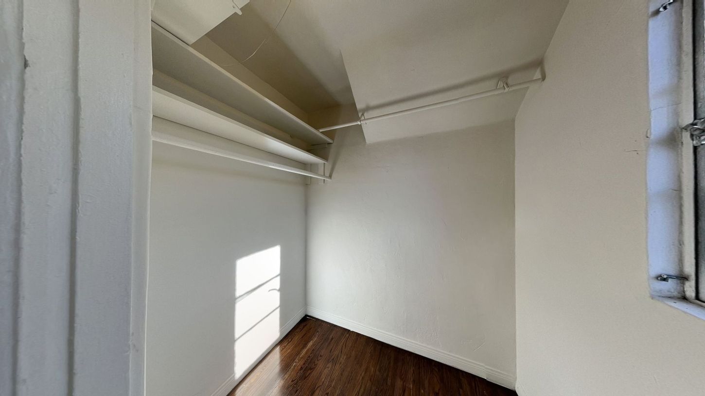 Empty white closet with a rod and shelves, hardwood floor, and window.