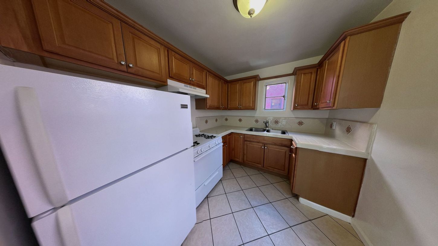 Kitchen with white appliances, brown cabinets, tiled floor, and small window.