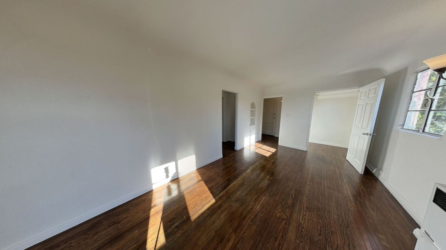 Empty room with hardwood floors, white walls, and bright sunlight streaming in.