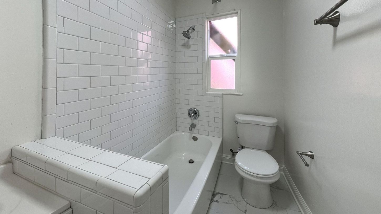 White-tiled bathroom with a bathtub, toilet, and window. Showerhead over the tub.