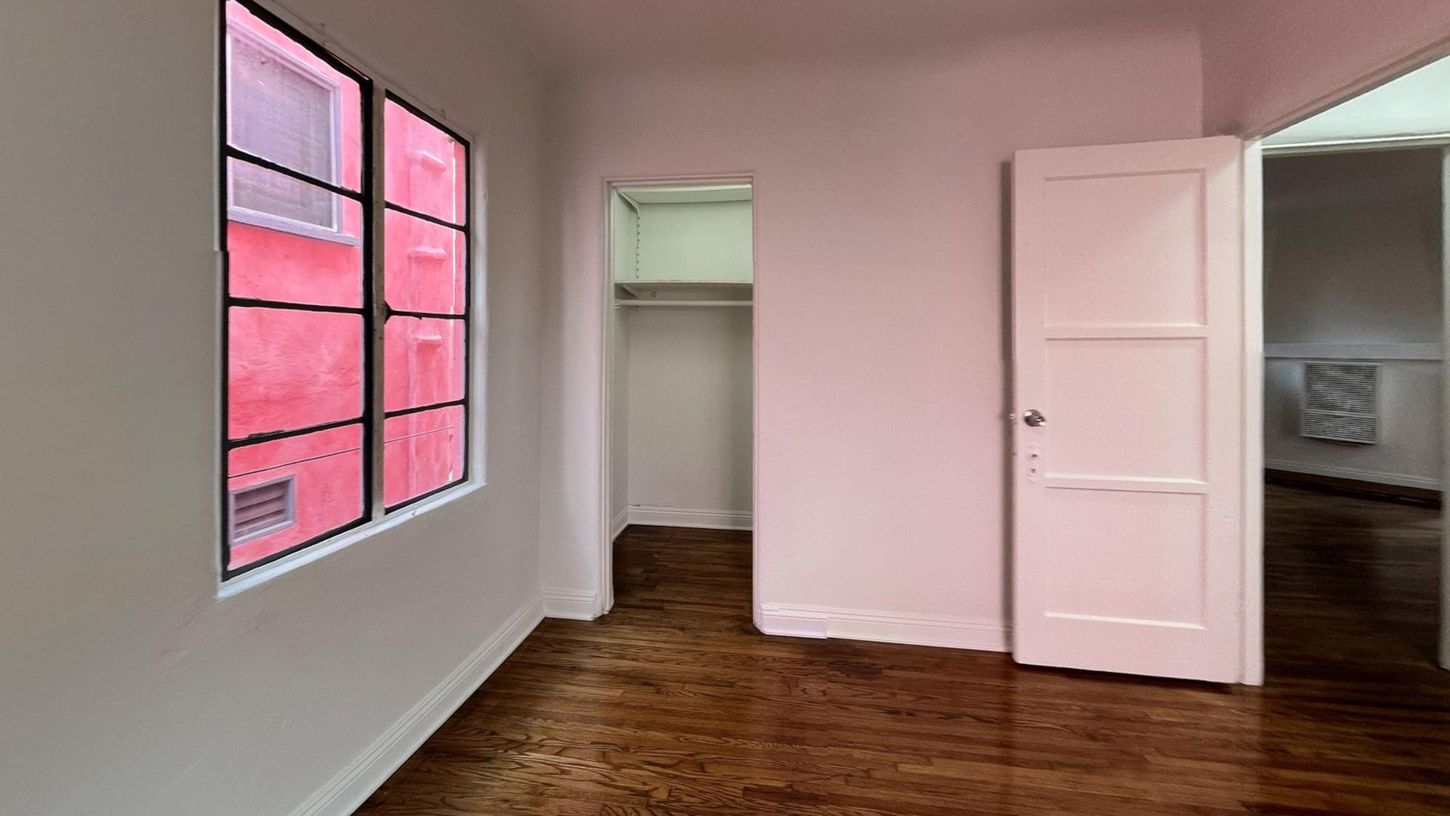 Empty room with a window, closet, and door. The walls are white, the floor is wood, and the window has a red exterior view.