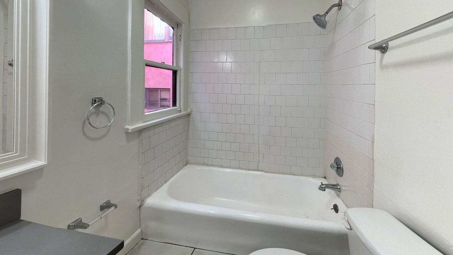 White bathroom with a tub, shower head, window, and toilet.