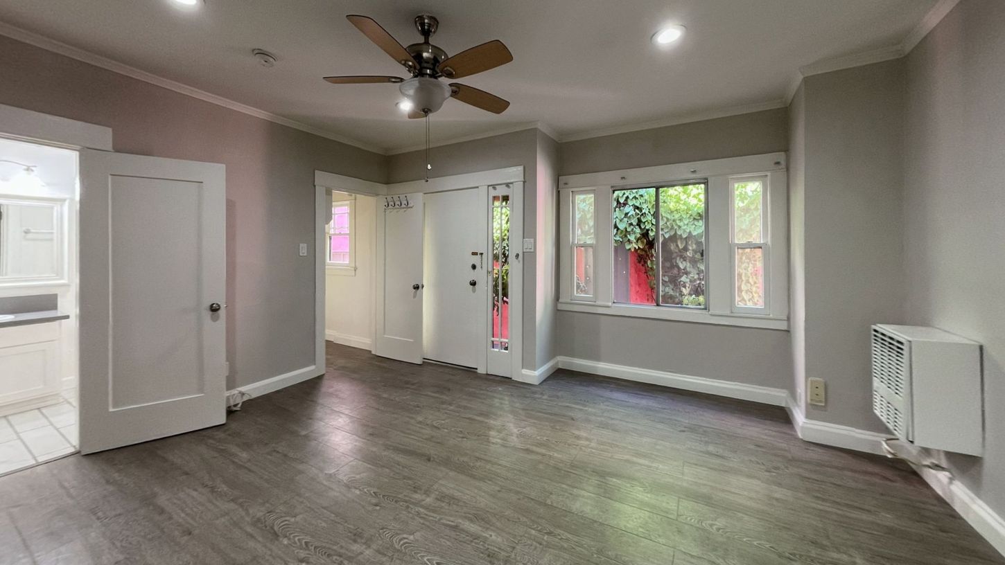Empty room with gray walls, dark wood-look flooring, white trim, ceiling fan, and window overlooking a small garden.