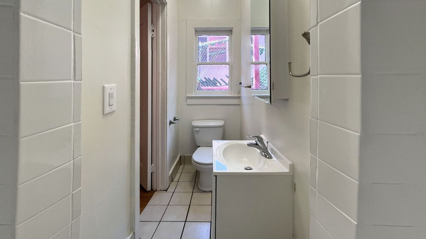 Narrow bathroom with white tile walls and floor, toilet, vanity, and a window.