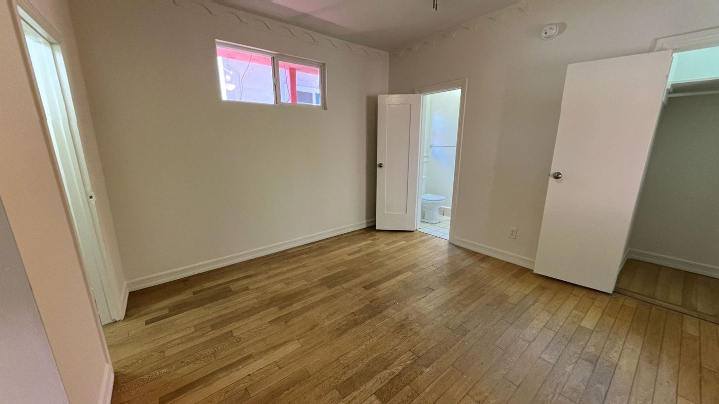 Empty room with wood floor, white walls, two open doors, and a small window.