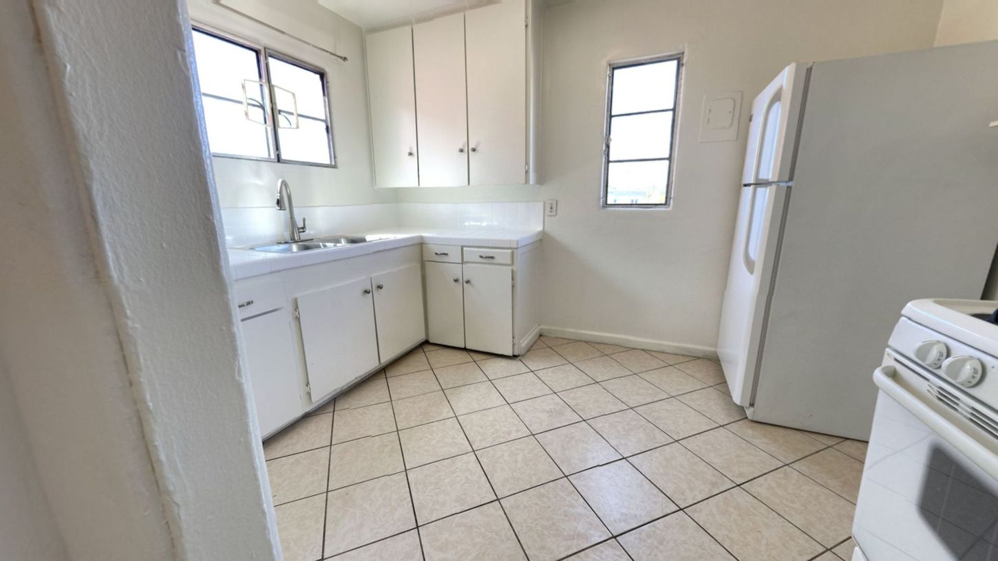 White kitchen with cabinets, sink, and refrigerator, tiled floor, two windows.