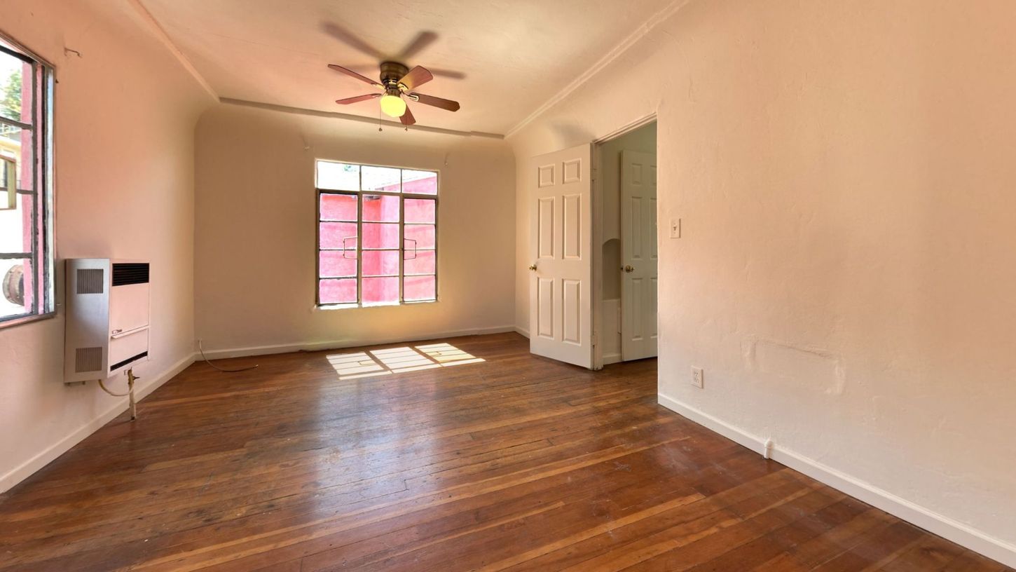 Empty room with hardwood floors, a ceiling fan, and a bright window with red trim.