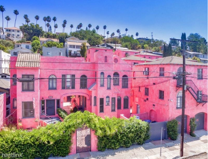 Pink stucco apartment buildings on a hillside, with palm trees and green foliage.