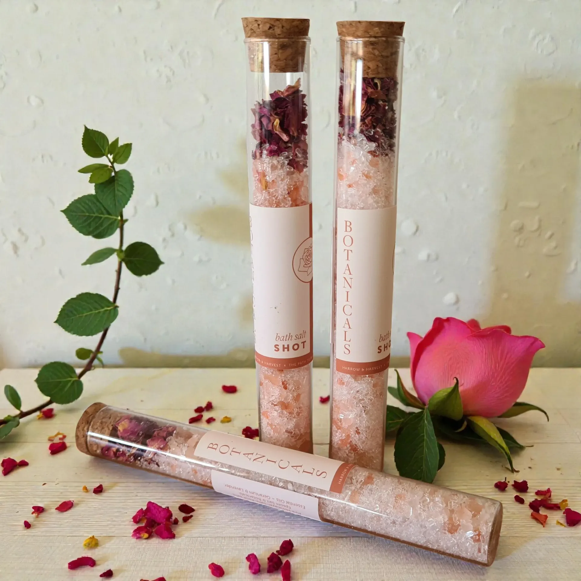 Three glass tubes of bath salts, dried rose petals, cork tops, labels, rose and leaves on table.