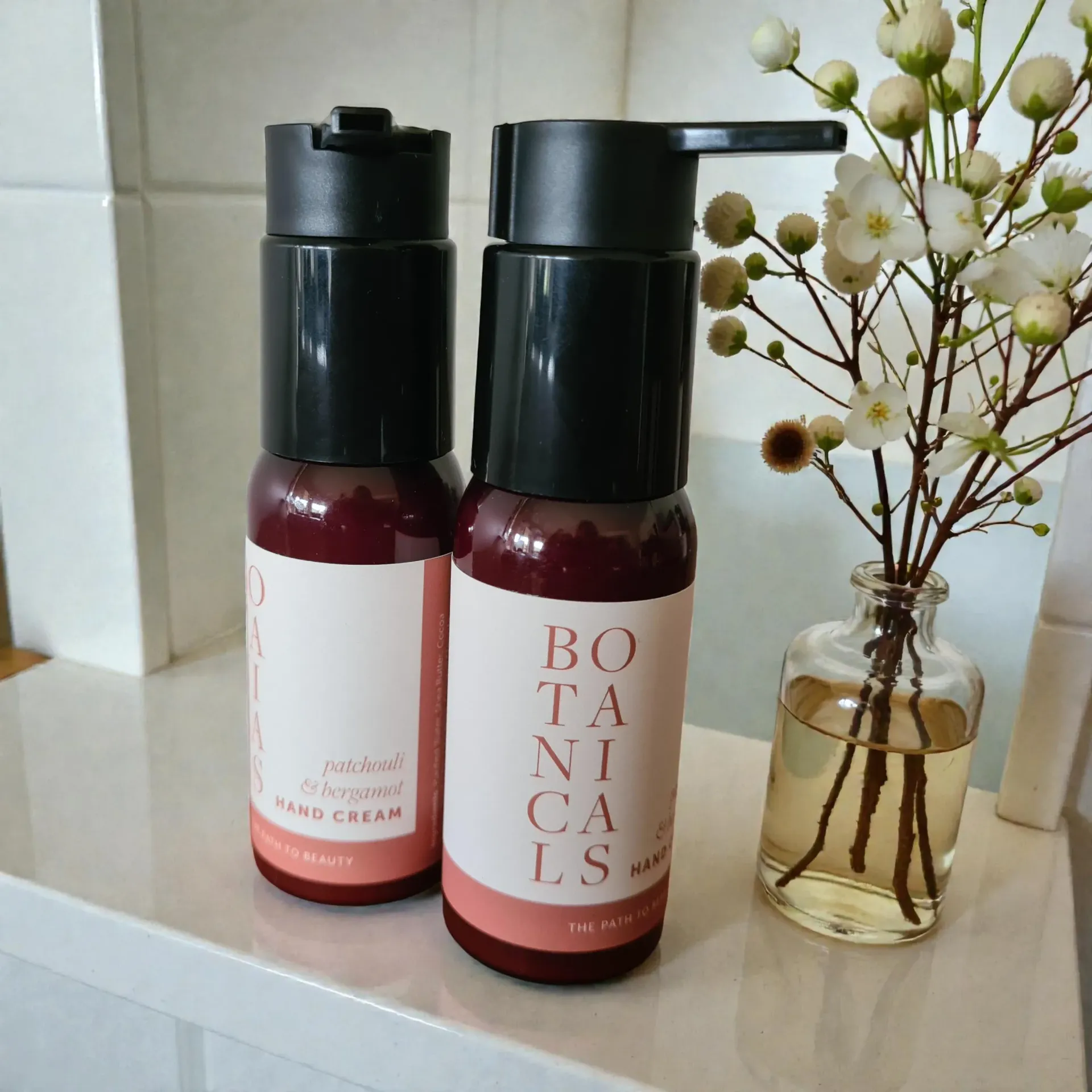 Two hand cream bottles, one with a pump, next to a small flower arrangement on a shelf.