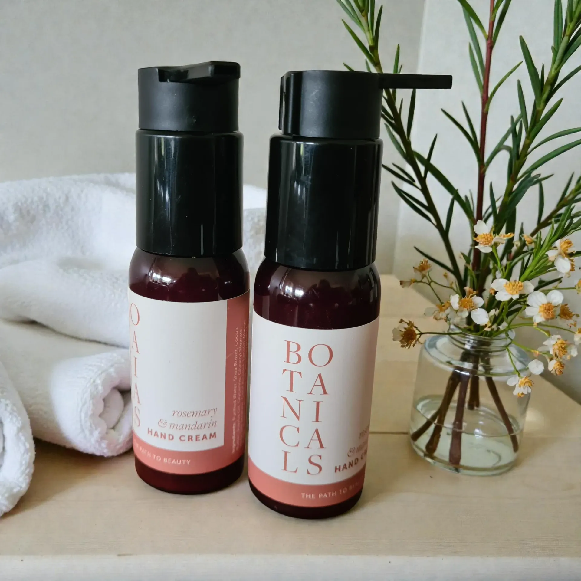 Two bottles of botanical hand cream with a white label, beside a glass vase and towels.