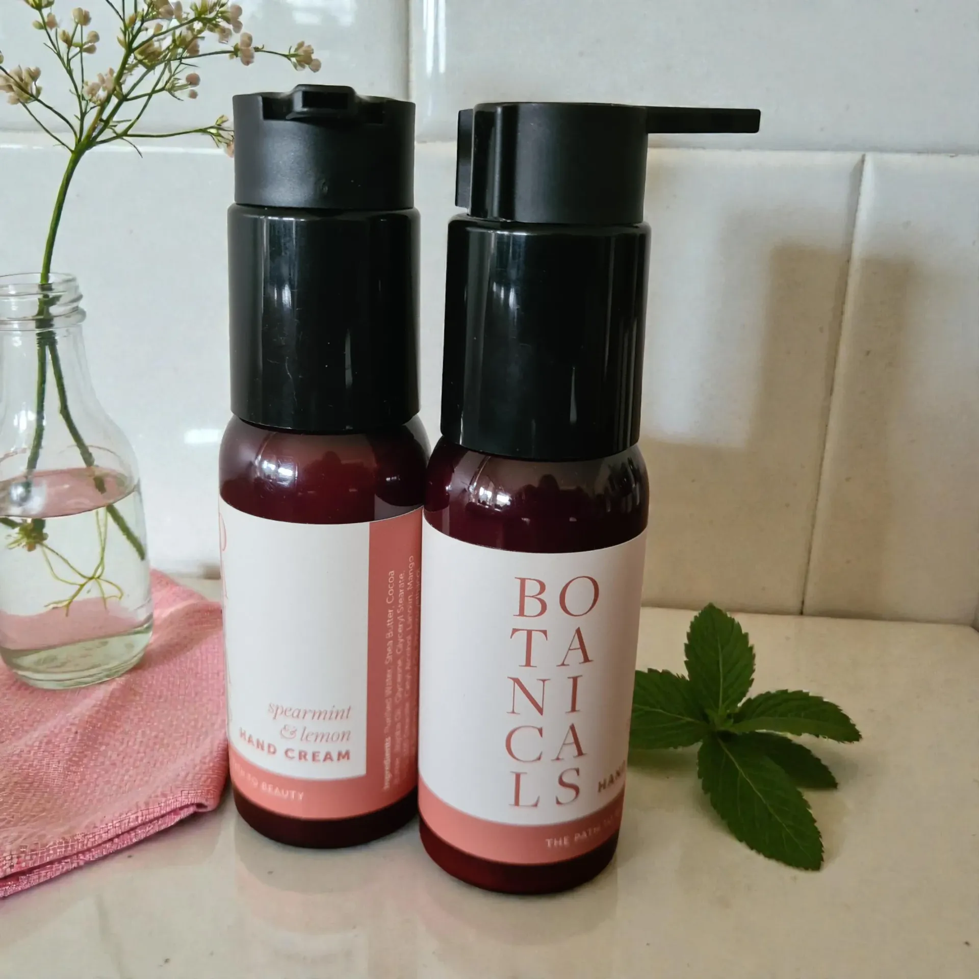 Two dark red bottles of botanical hand cream with black pumps, set on a white tiled surface.