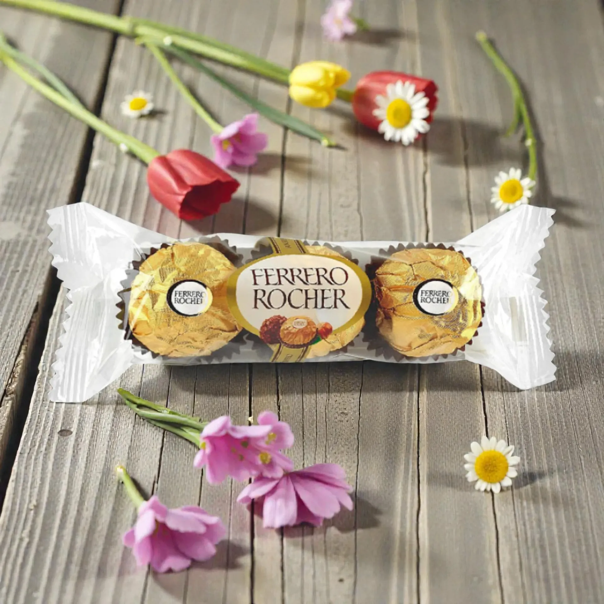 Ferrero Rocher candies in wrapper, next to tulips and daisies on wood.