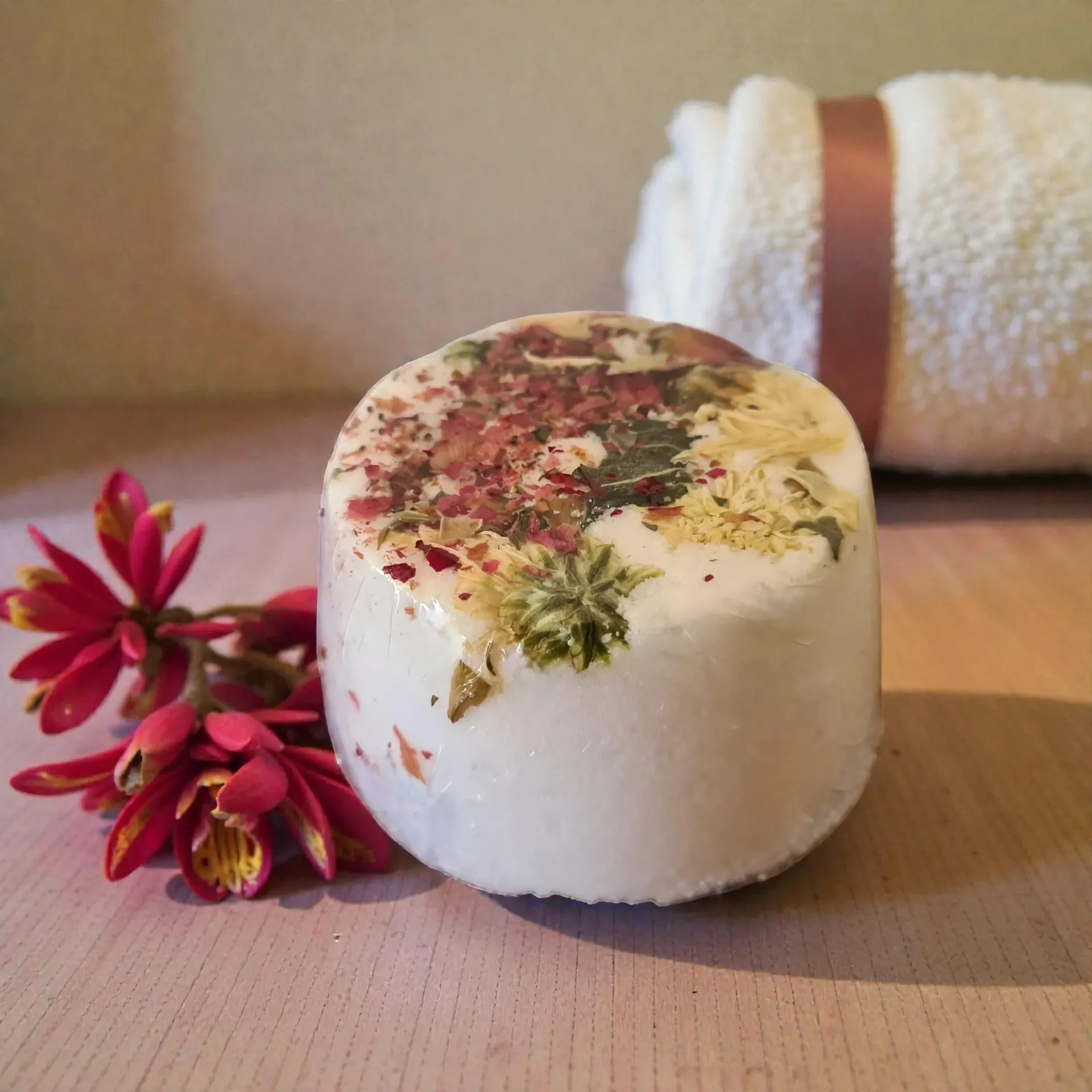 A white bath bomb with dried flowers, a red flower, and rolled towel with brown ribbon.