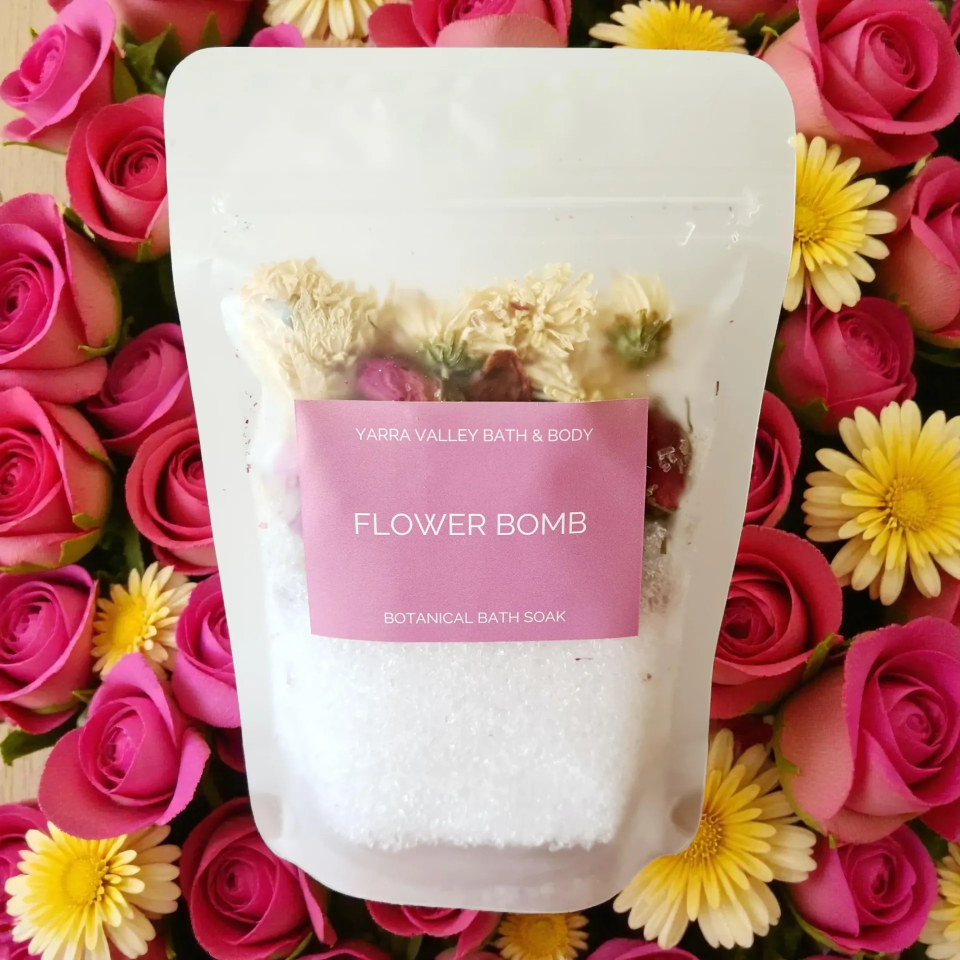Bag of bath salts with dried flowers, surrounded by pink roses and yellow flowers.