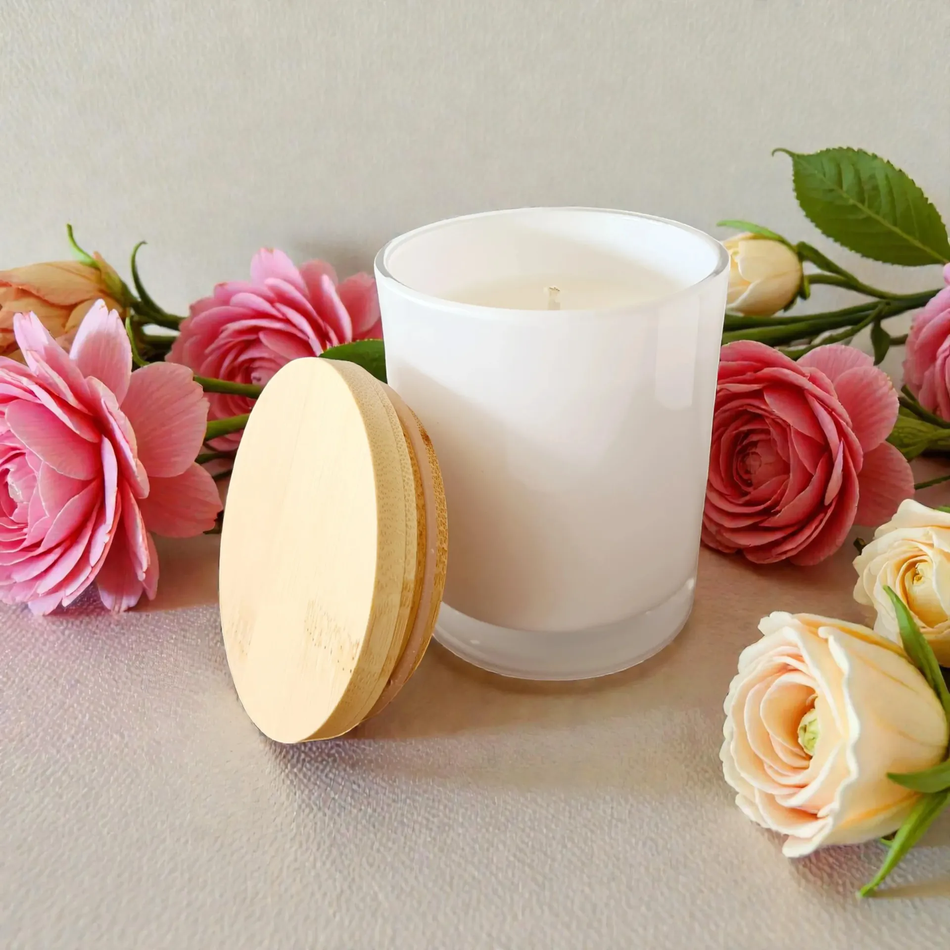 White candle in glass jar with wooden lid, surrounded by pink and cream roses on a silver surface.
