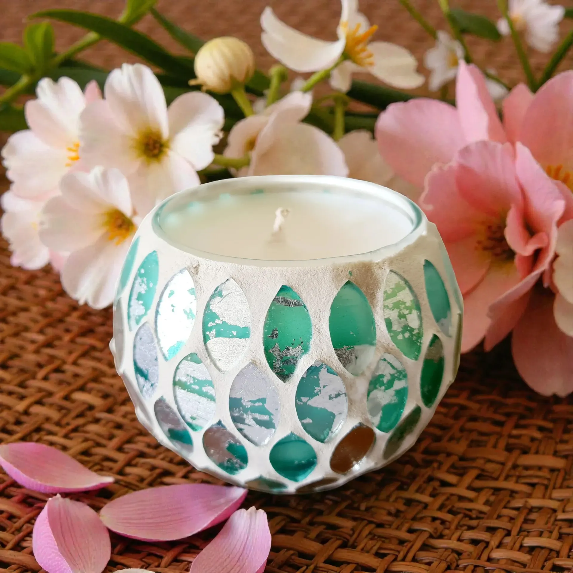 Mosaic glass candle in front of pink and white flowers on a woven surface.