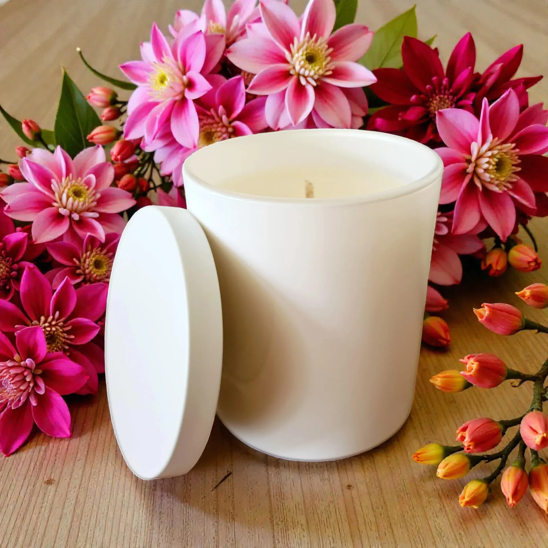 White candle with lid, surrounded by pink flowers on a wooden surface.