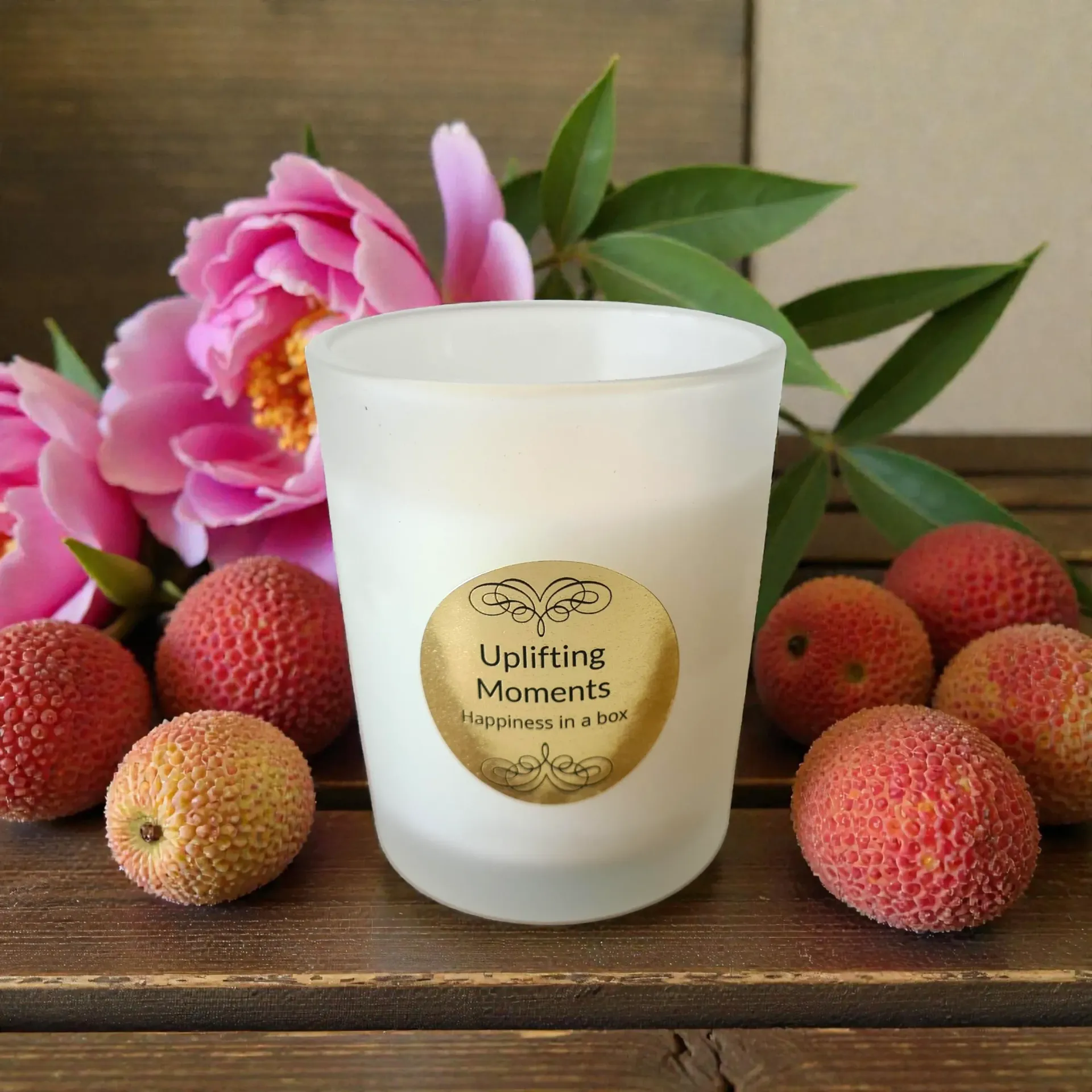 Candle in frosted glass with gold label, surrounded by lychees and pink peonies on wood.