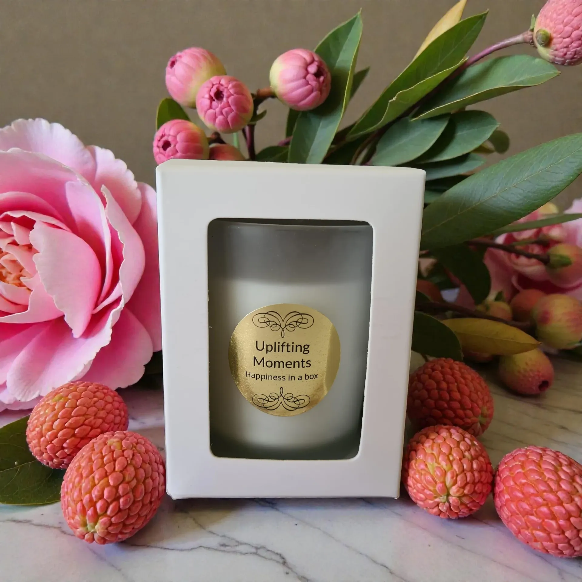 Candle in frosted glass with gold label, surrounded by lychees and pink peonies on wood.