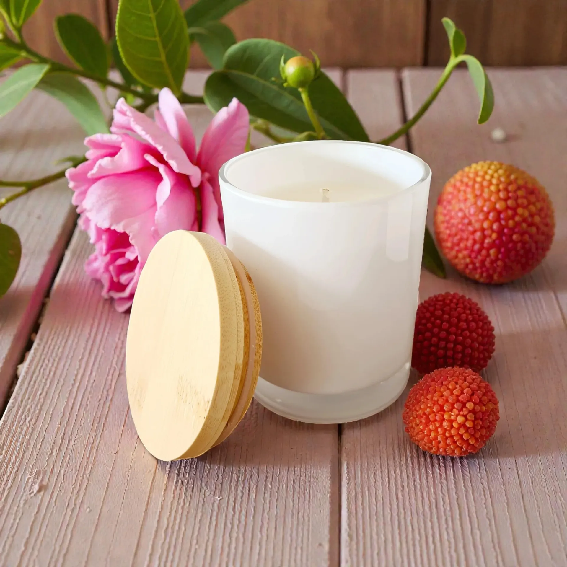 White candle in glass with pink flower, lychees, and macaron on a pink wooden surface.
