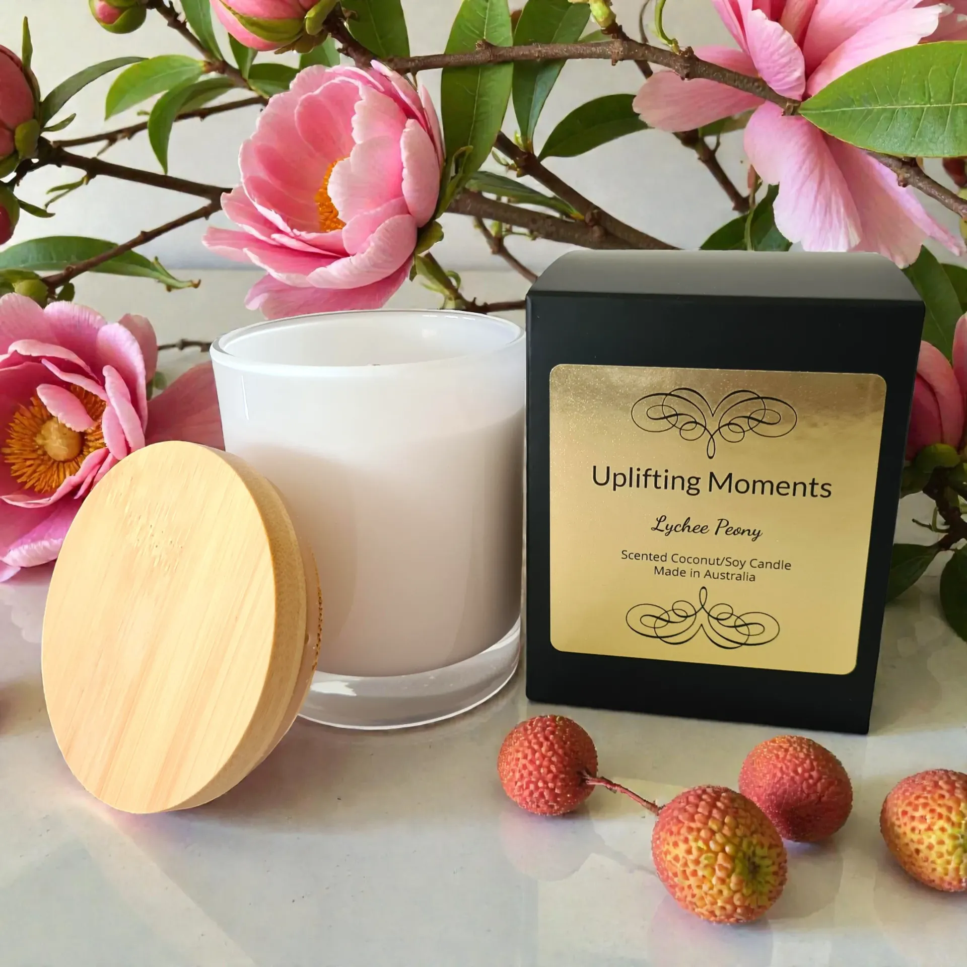 White candle in glass with pink flower, lychees, and macaron on a pink wooden surface.