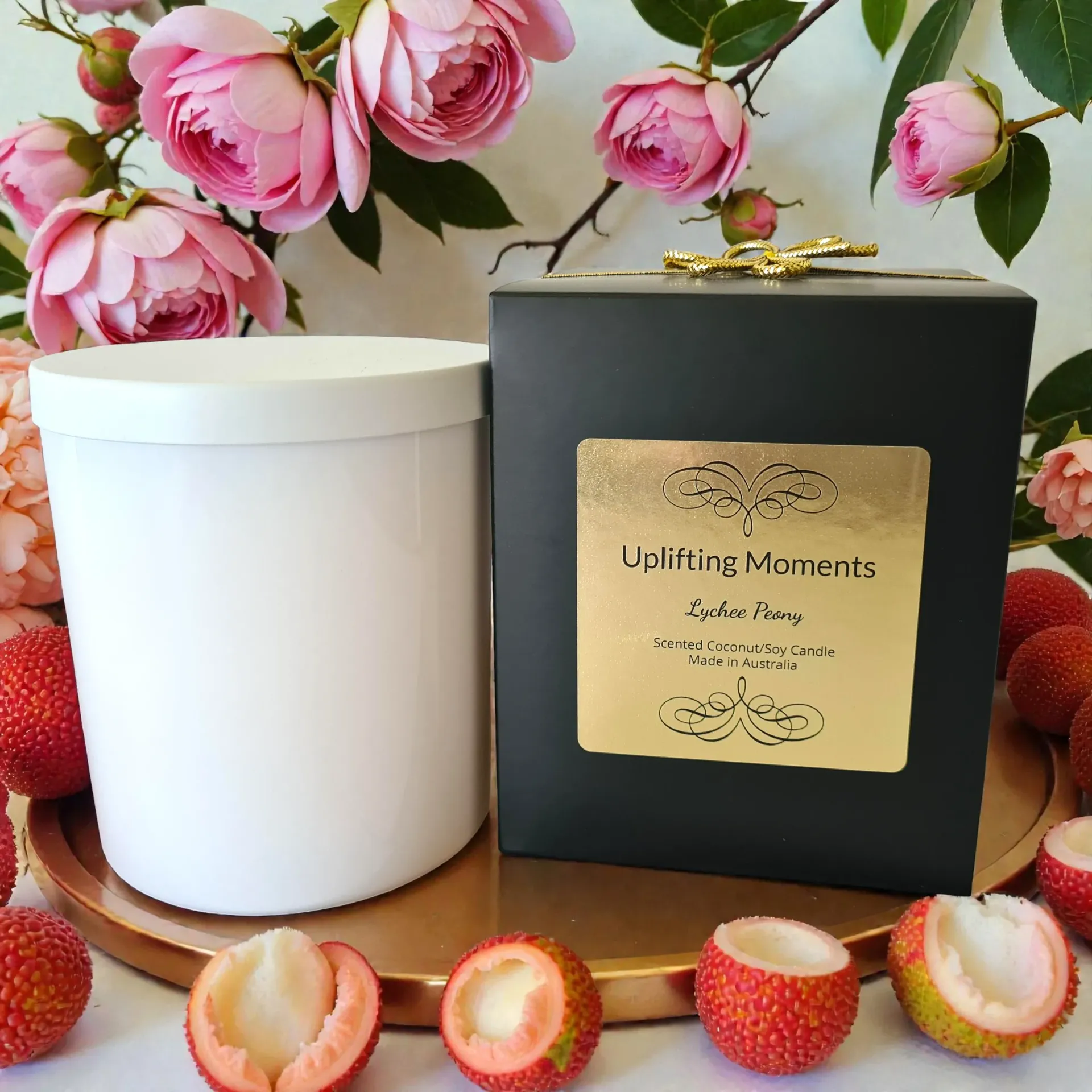 White candle with lid, pink peonies, and lychees on wooden surface.