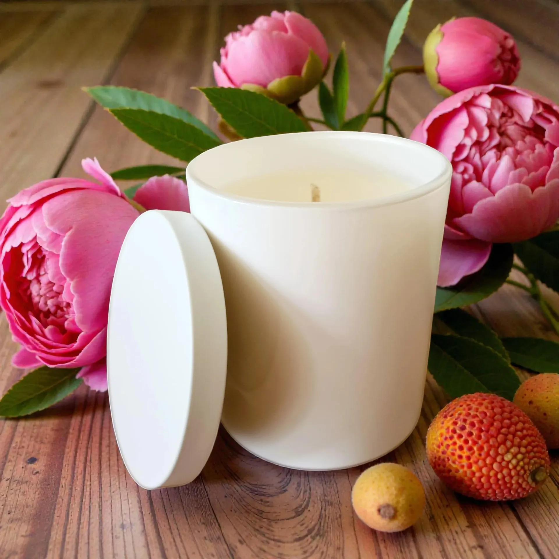 White candle with lid, pink peonies, and lychees on wooden surface.