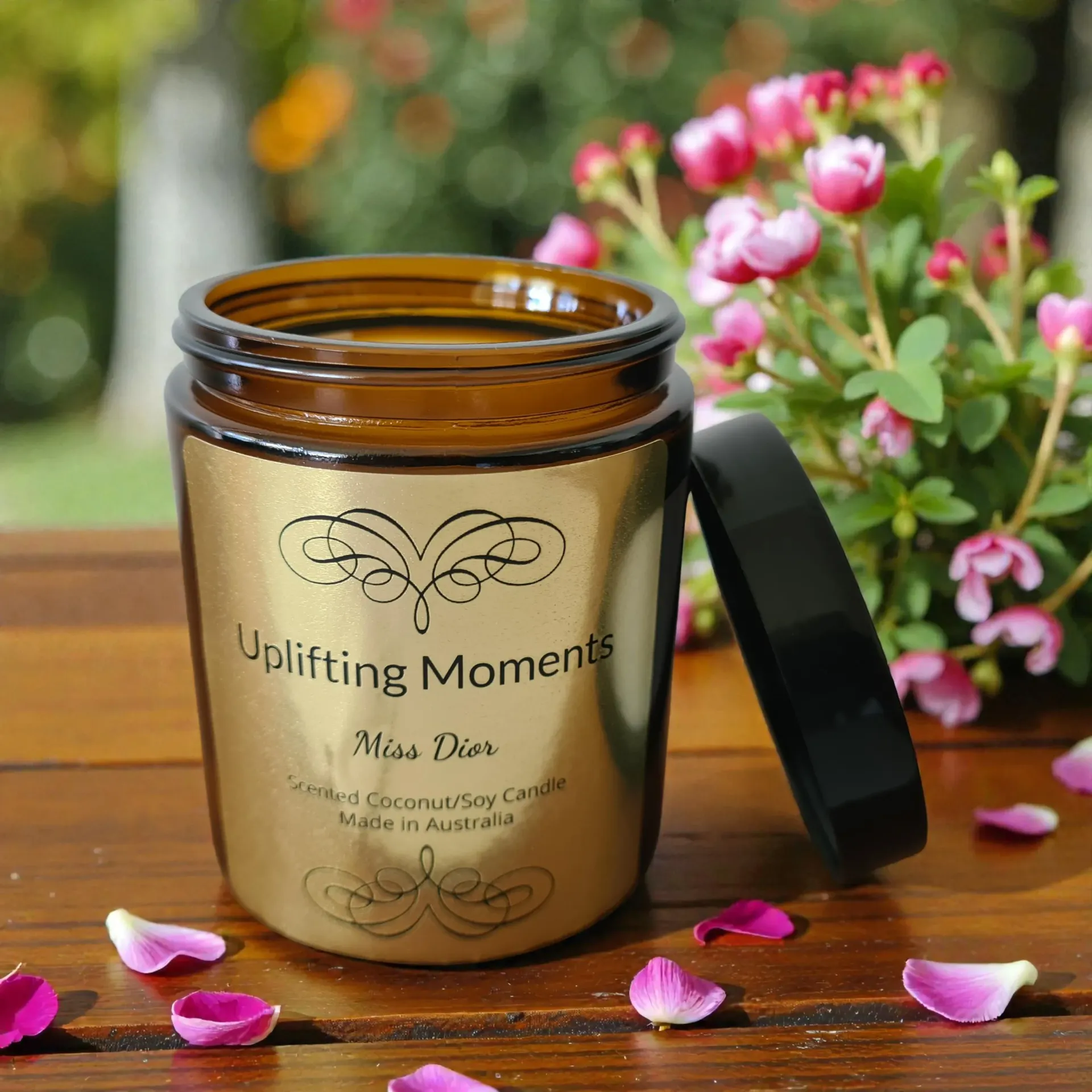 Gold candle with black lid, “Uplifting Moments” label, surrounded by pink flowers.