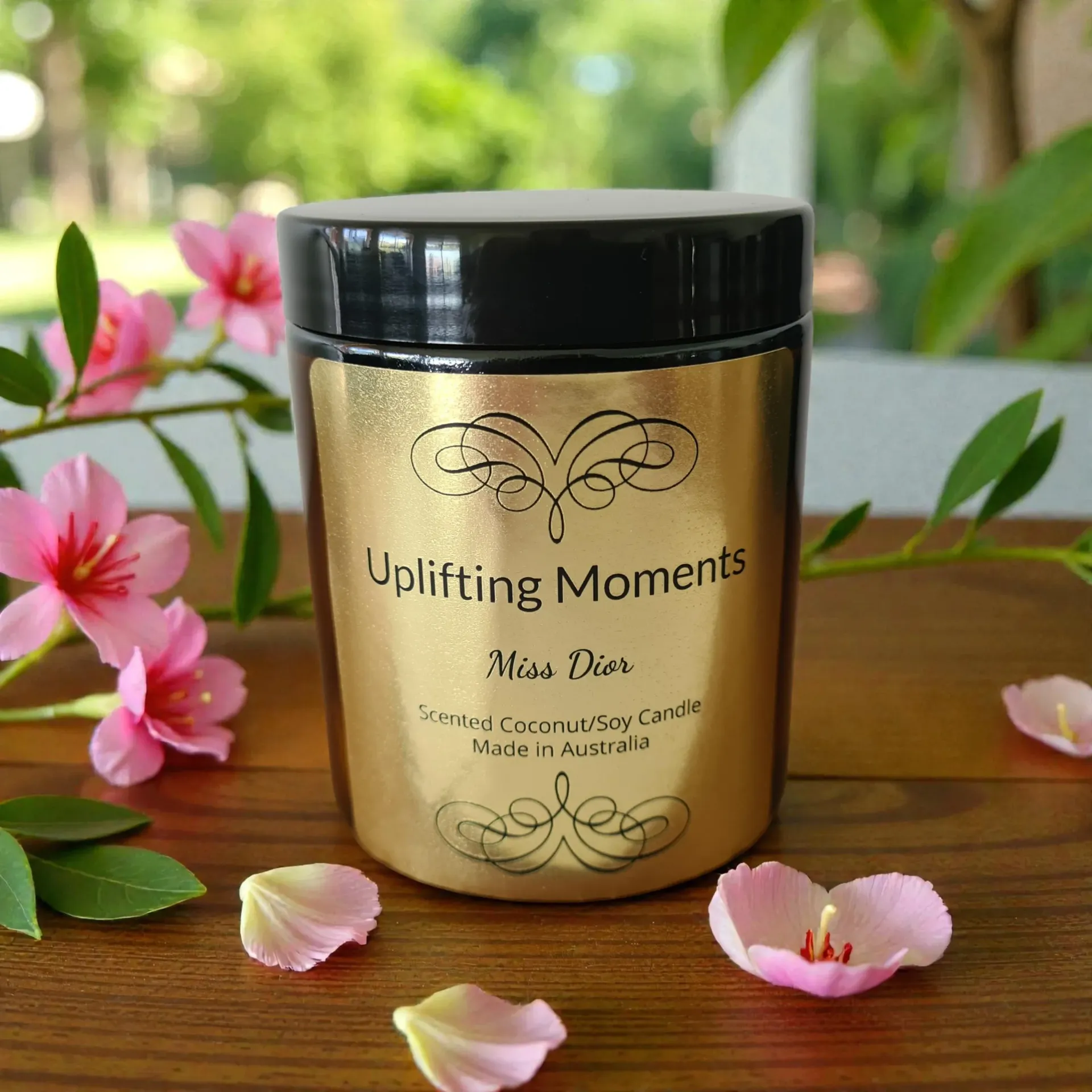 Gold candle with black lid, “Uplifting Moments” label, surrounded by pink flowers.