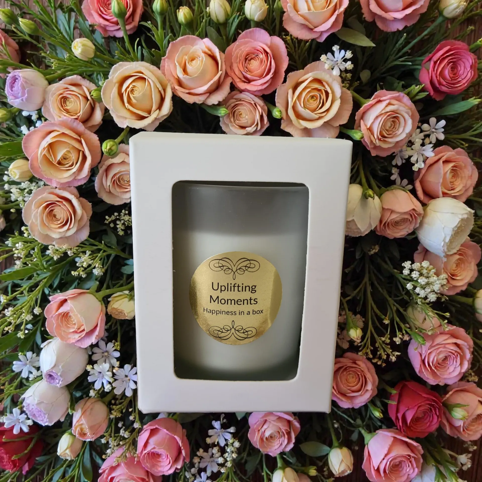 Frosted white candle with gold label, surrounded by colorful roses and greenery on wooden surface.