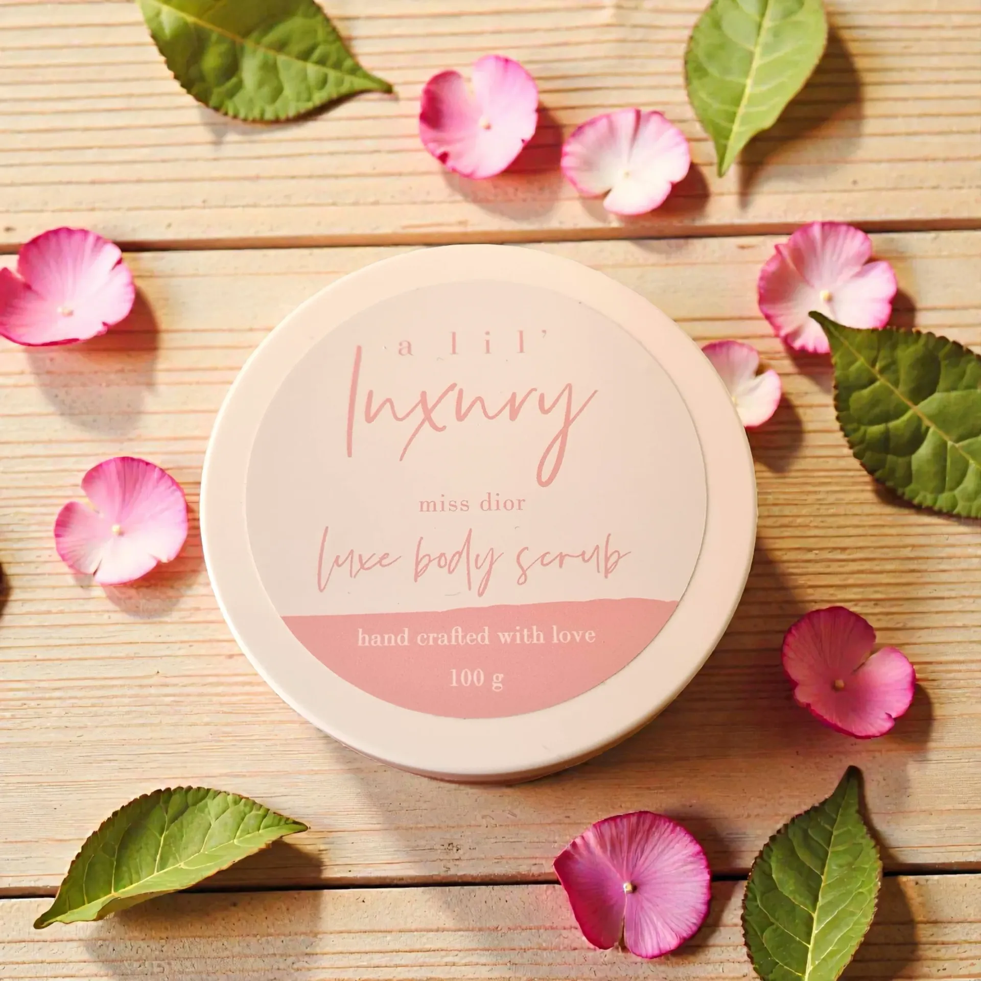 Pink body scrub jar with rose petals on a wooden surface.