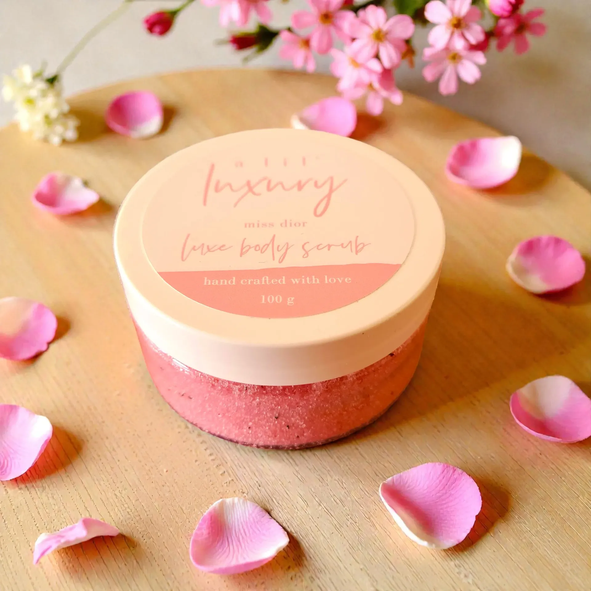 Pink body scrub jar with rose petals on a wooden surface.