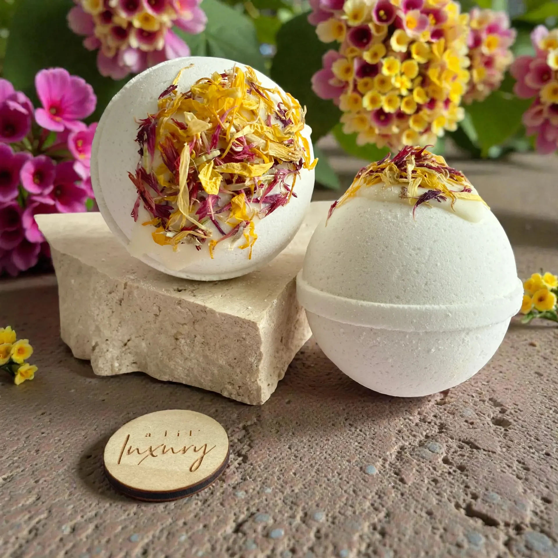 Two white bath bombs with floral accents, on a stone, flowers in background.