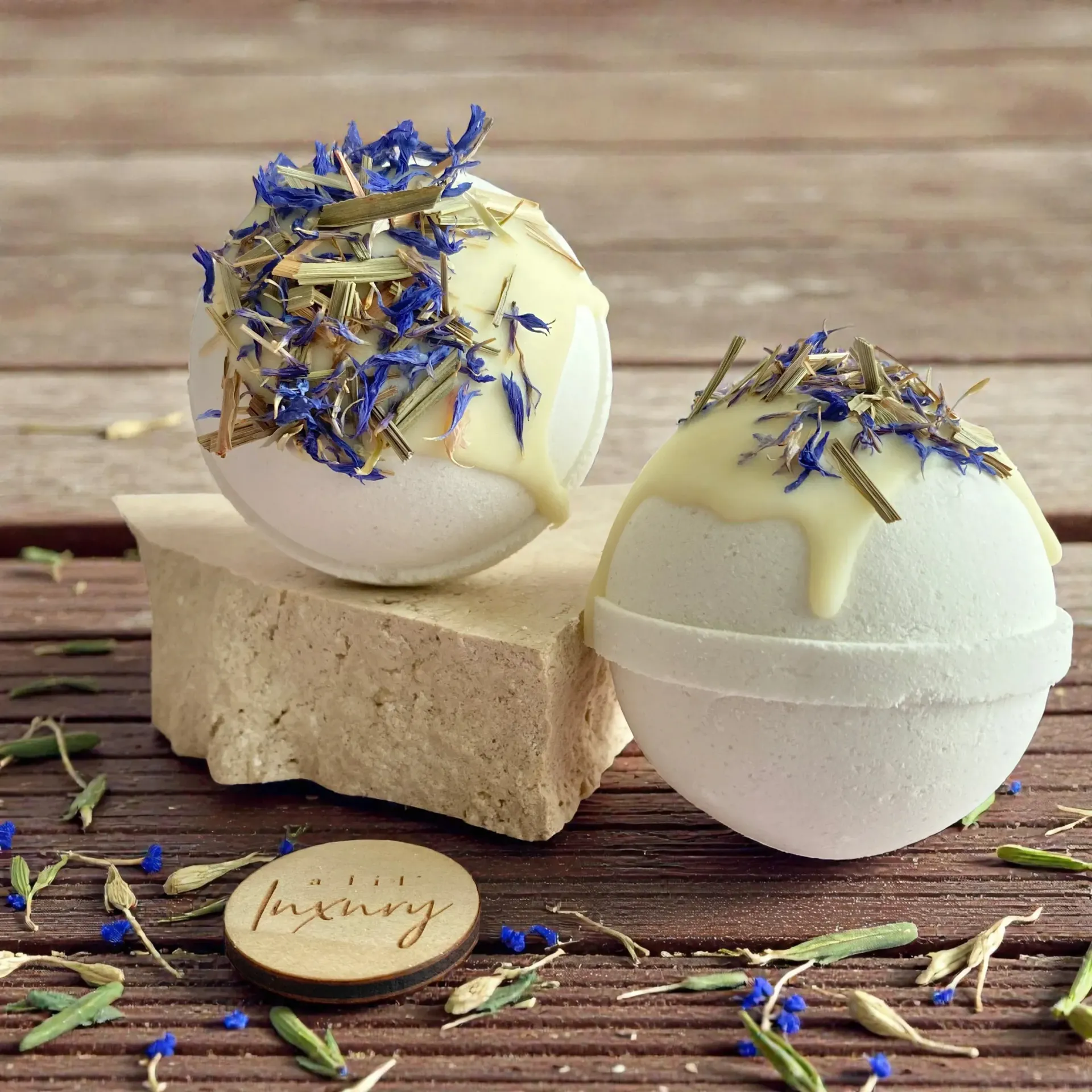 Two white bath bombs with yellow drizzled tops, blue flower petals, and wooden coaster.