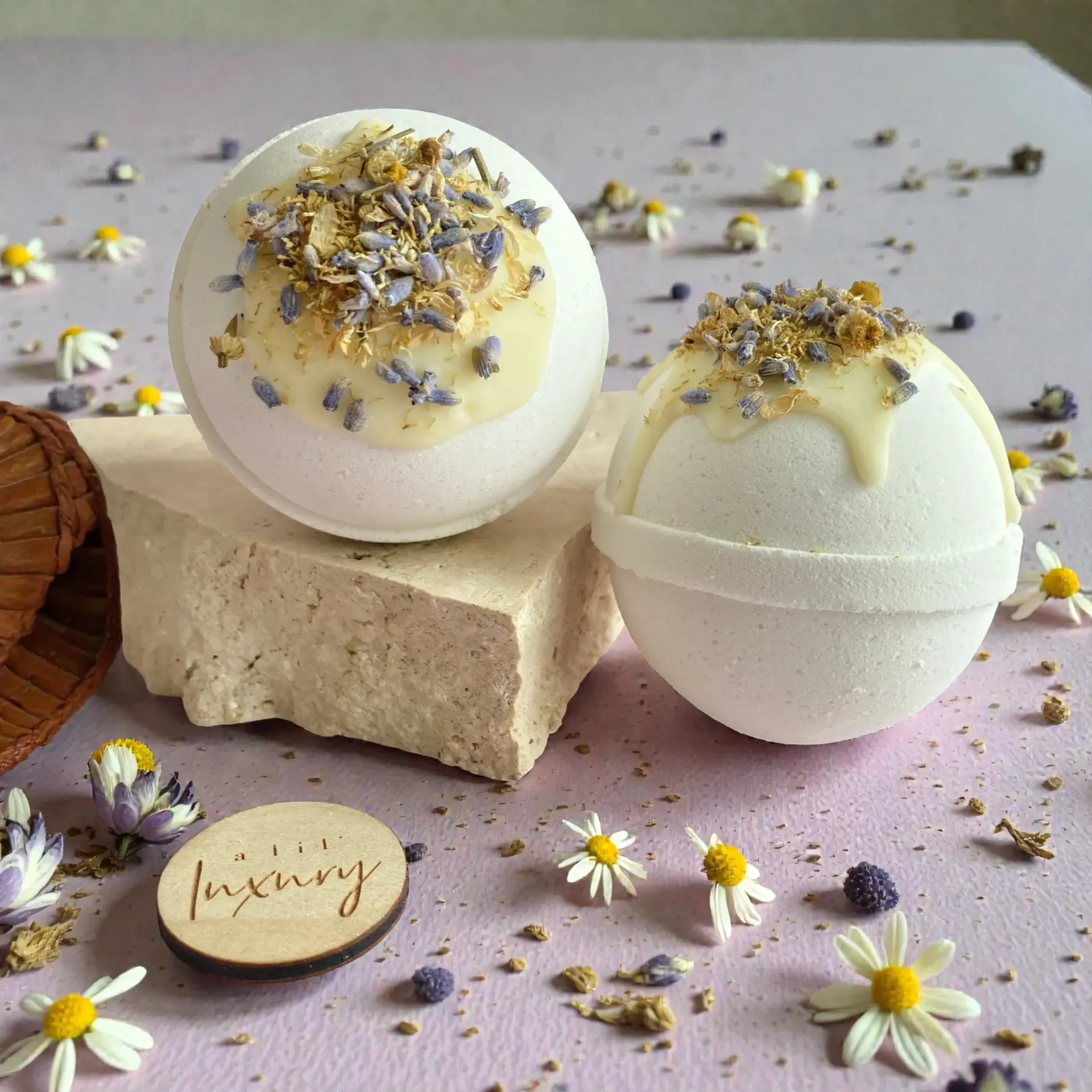 Two white bath bombs with floral toppings, on a stone block, scattered flowers, and a wooden label.