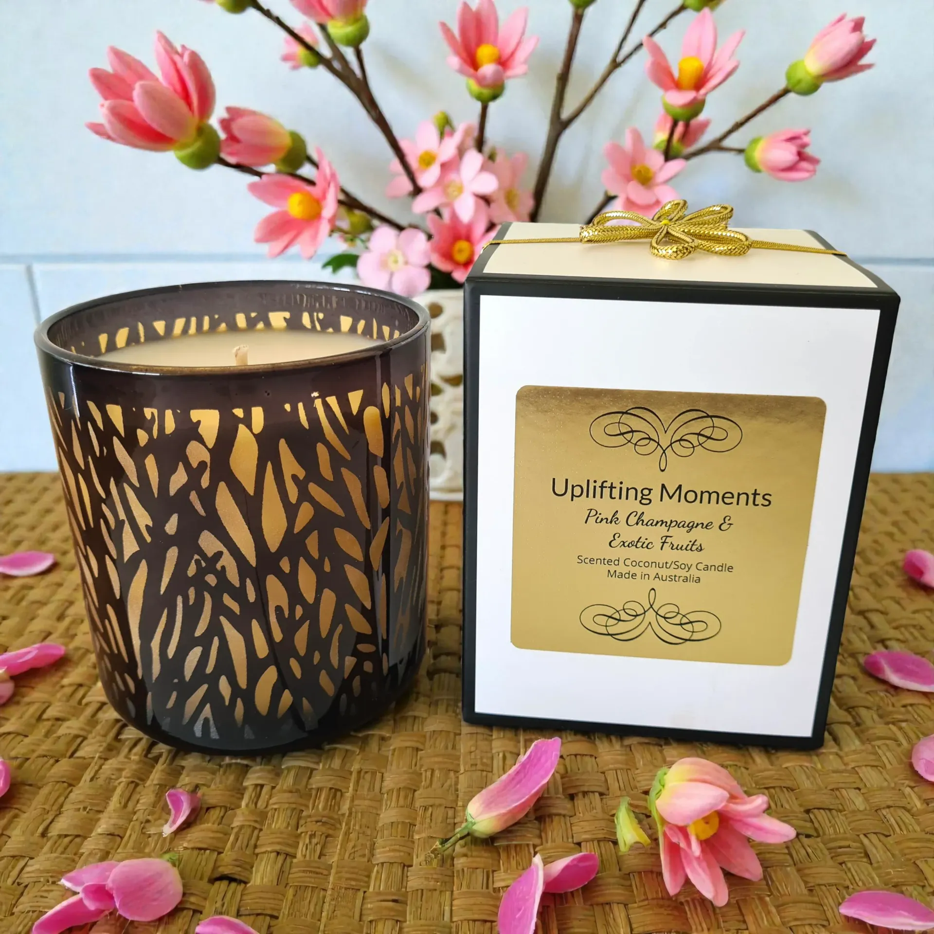 Candle in a dark, patterned glass jar, next to a rose and wine glass, on a table.