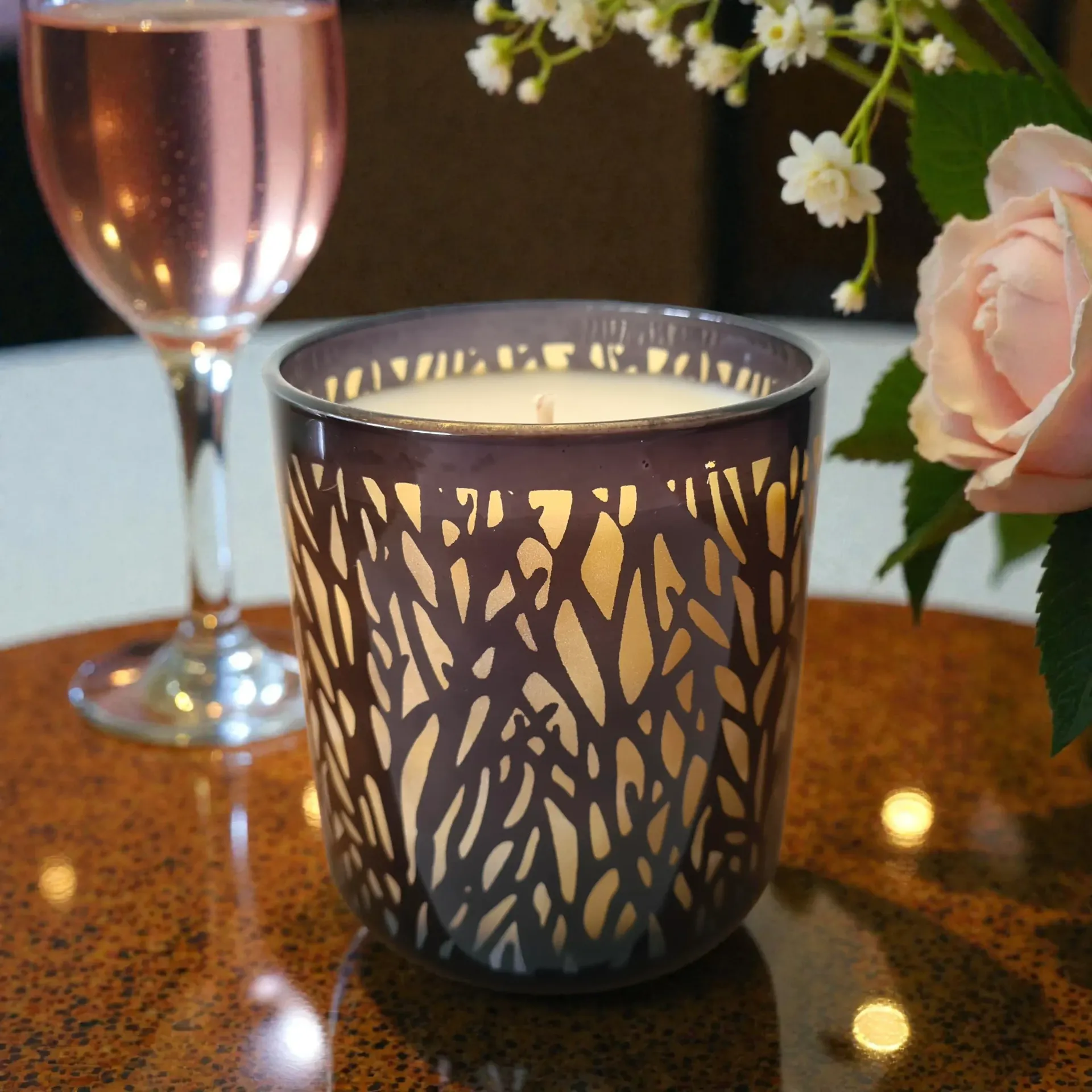 Candle in a dark, patterned glass jar, next to a rose and wine glass, on a table.