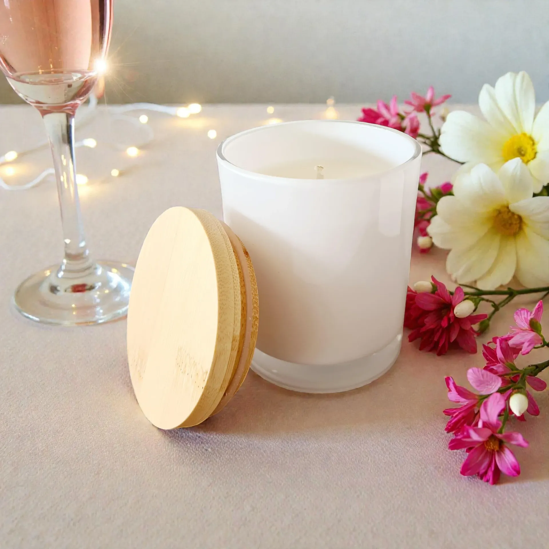 White candle next to its wooden lid, wine glass, flowers and string lights.