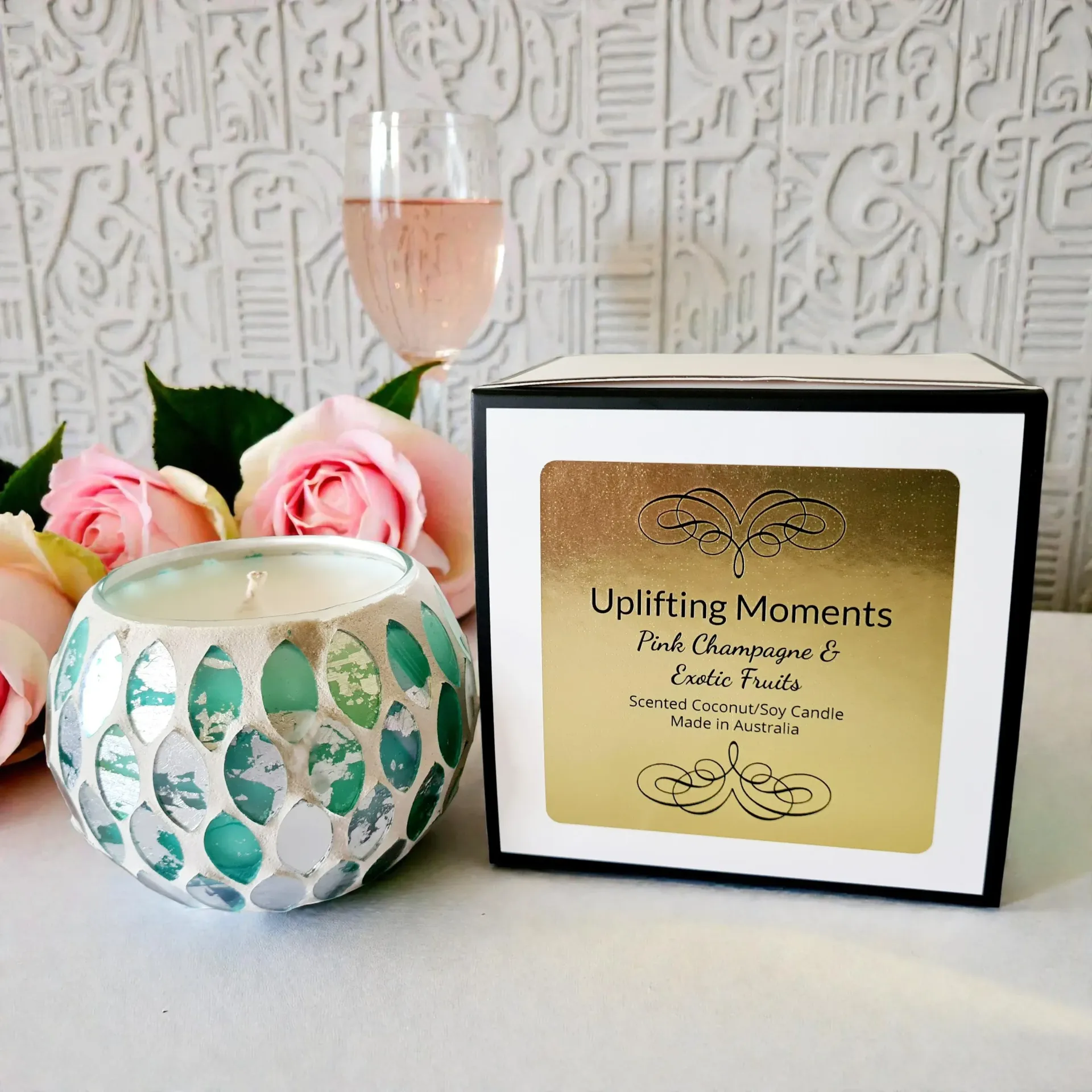 Candle in mosaic glass bowl, surrounded by flowers, with a glass of wine in the background.