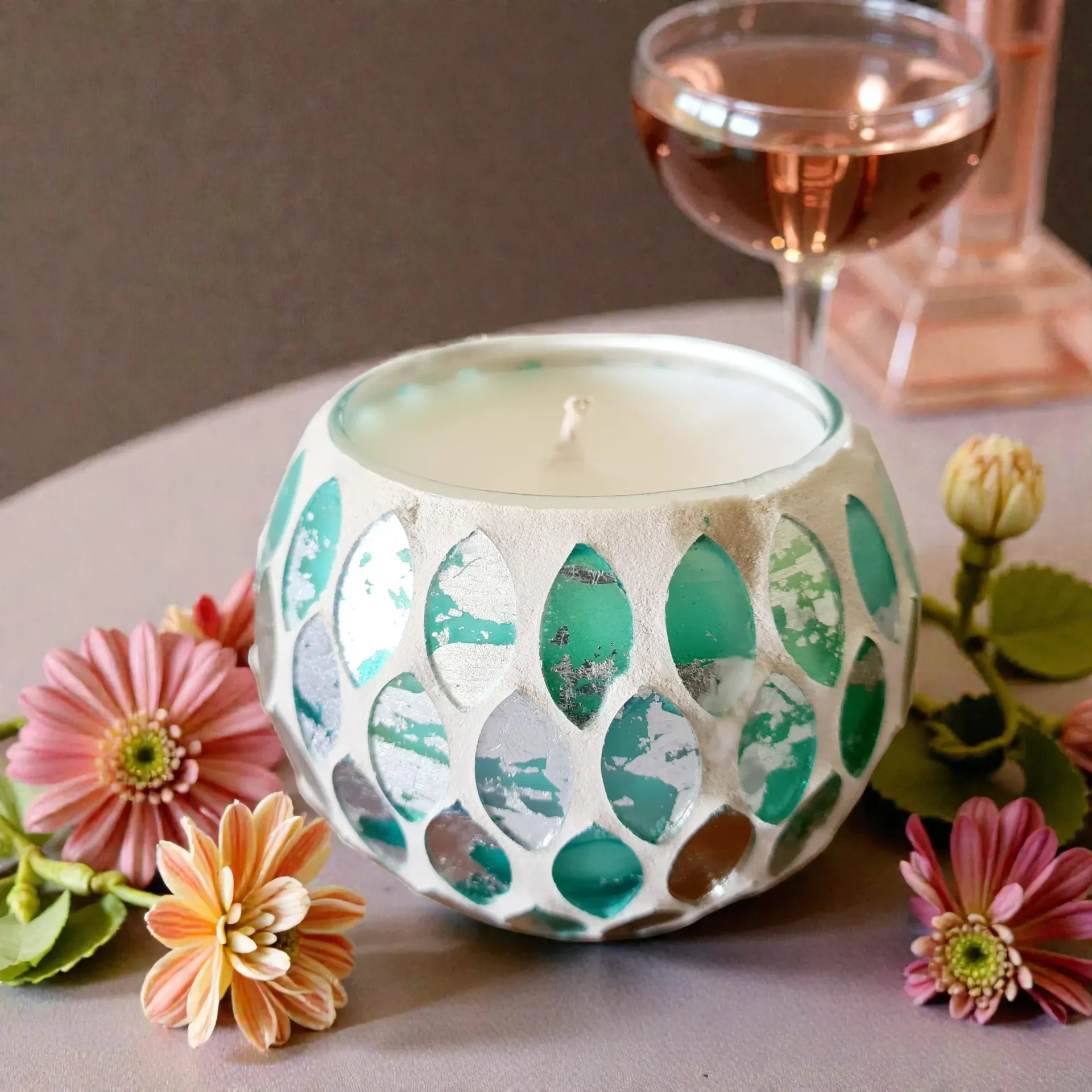 Candle in mosaic glass bowl, surrounded by flowers, with a glass of wine in the background.
