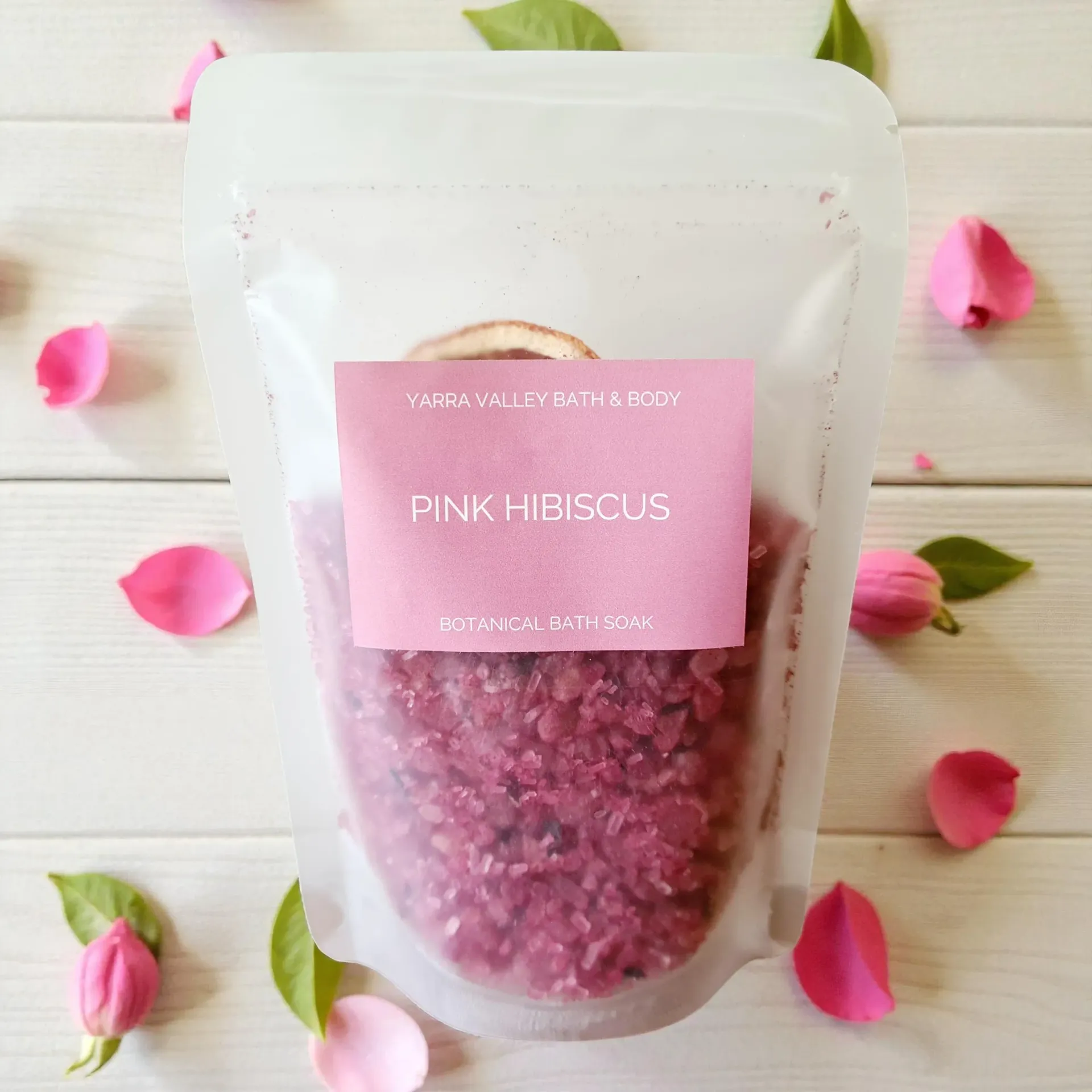 Bag of pink hibiscus bath salts with rose petals and leaves on a white wooden surface.