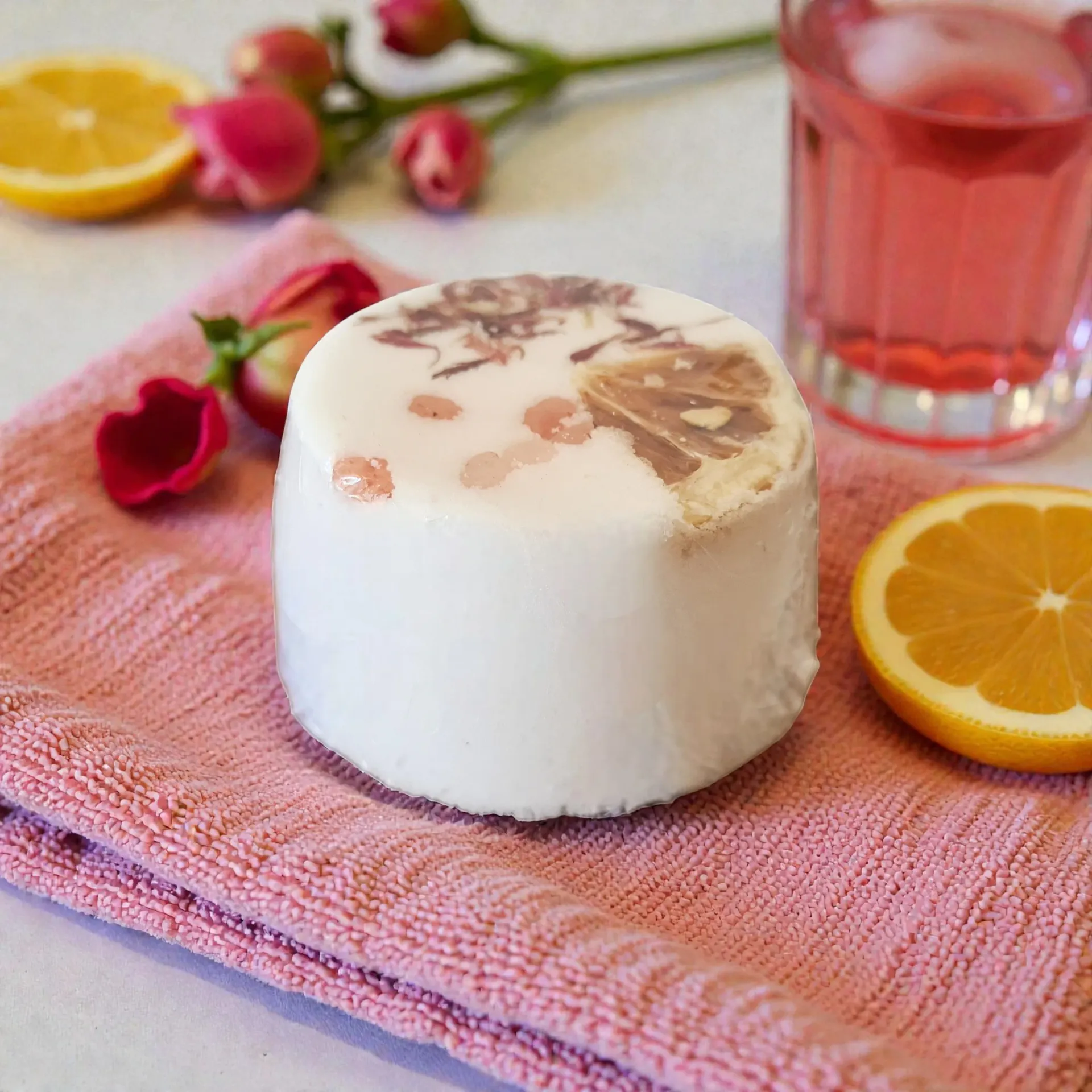 A white soap with lemon and rose decorations, on a pink towel, with lemon slices and pink drink.