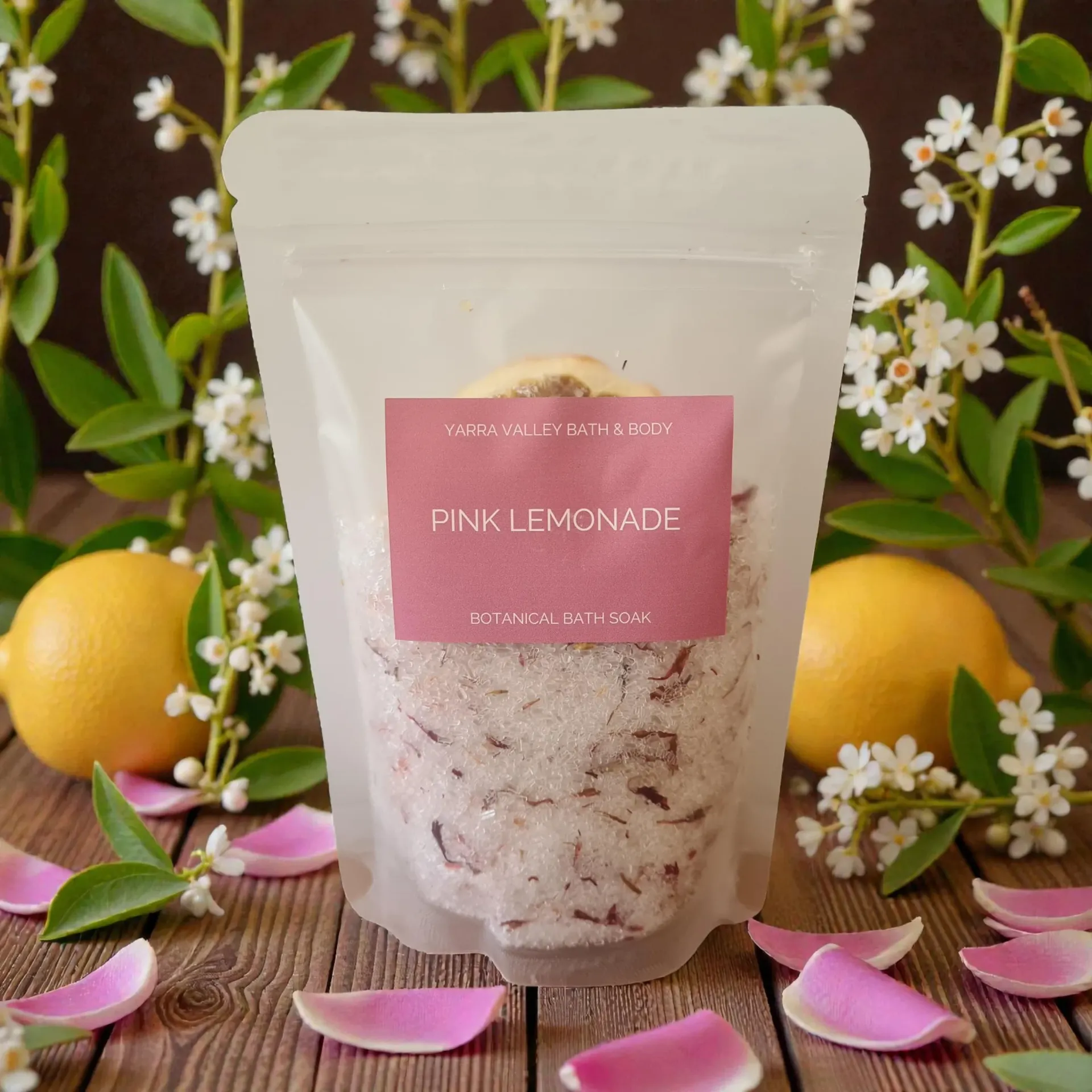 Pink lemonade bath soak in a clear pouch, lemons, flowers, pink petals on wooden surface.