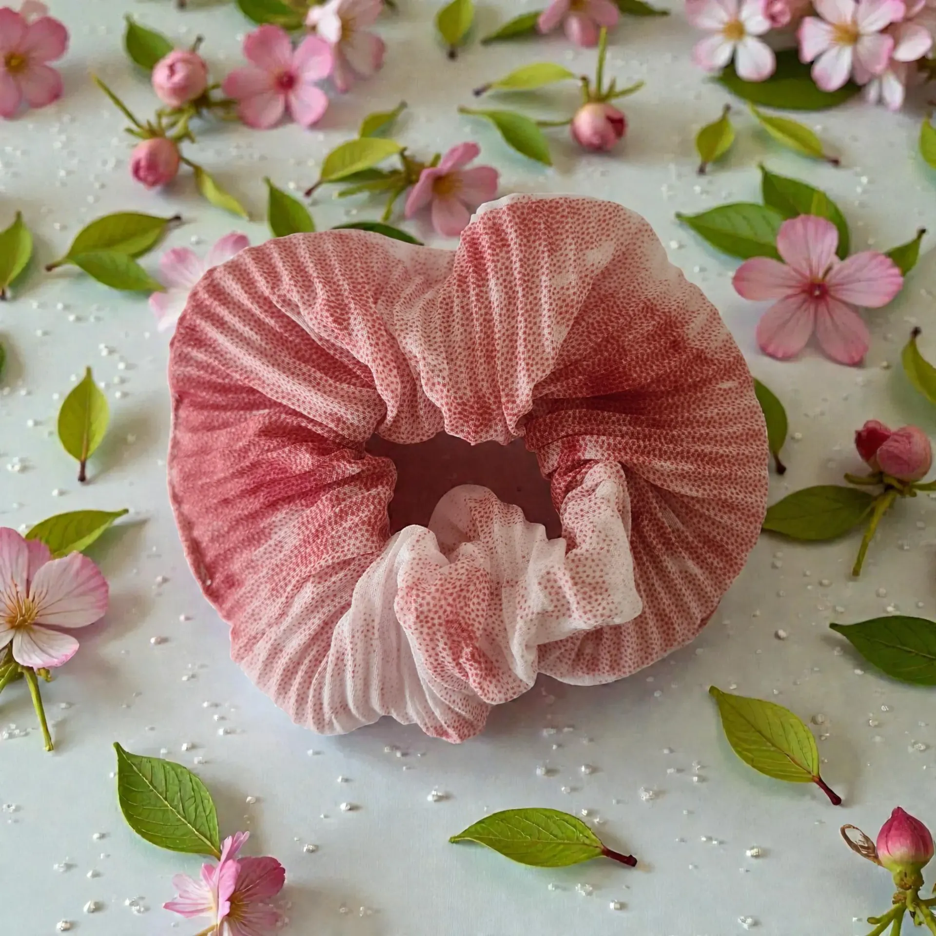Pink and white scrunchie surrounded by pink flowers and green leaves on a white surface.
