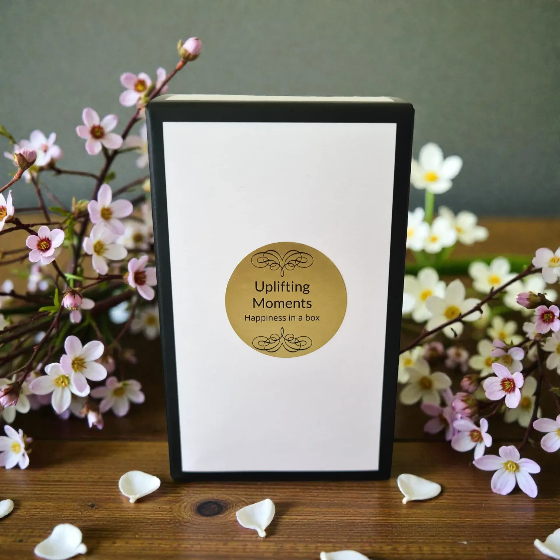 White candle in a frosted glass with a gold label, surrounded by flowers on a wooden table, outside with green plants.