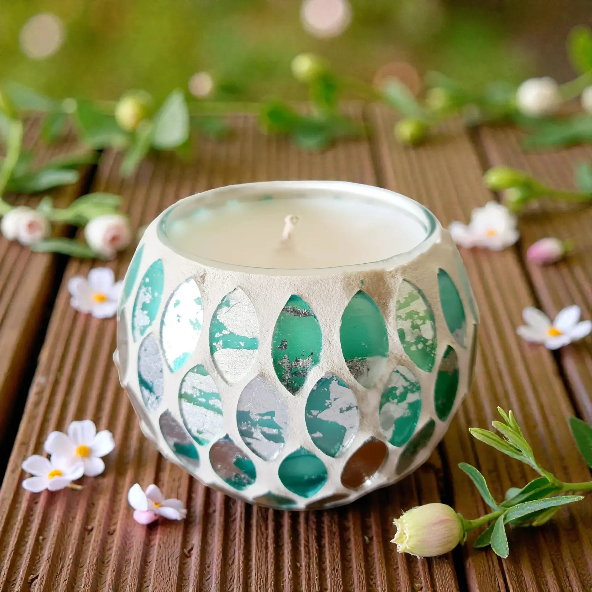 Decorative candle in a mosaic glass holder on a wooden surface, with flowers and greenery.