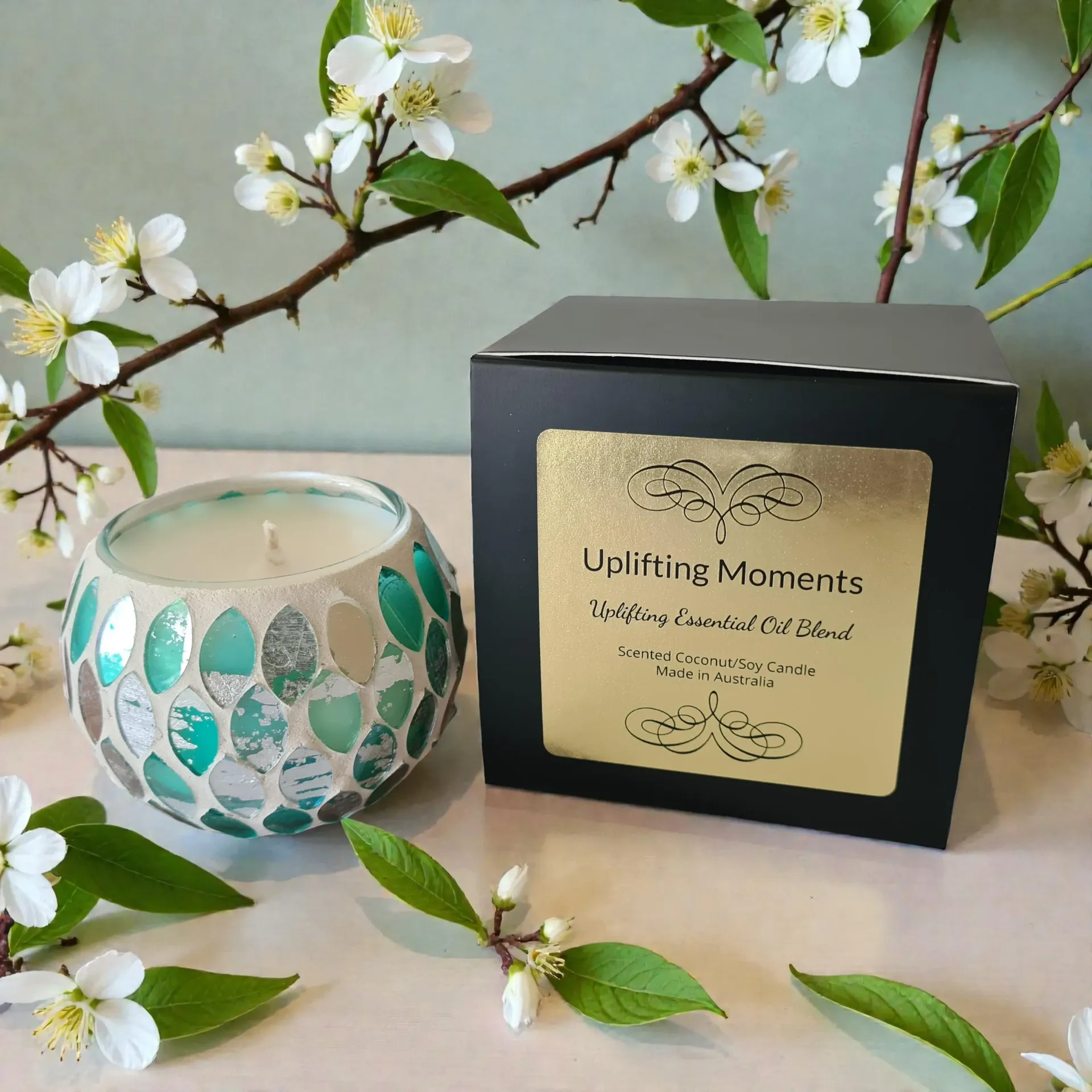 Decorative candle in a mosaic glass holder on a wooden surface, with flowers and greenery.